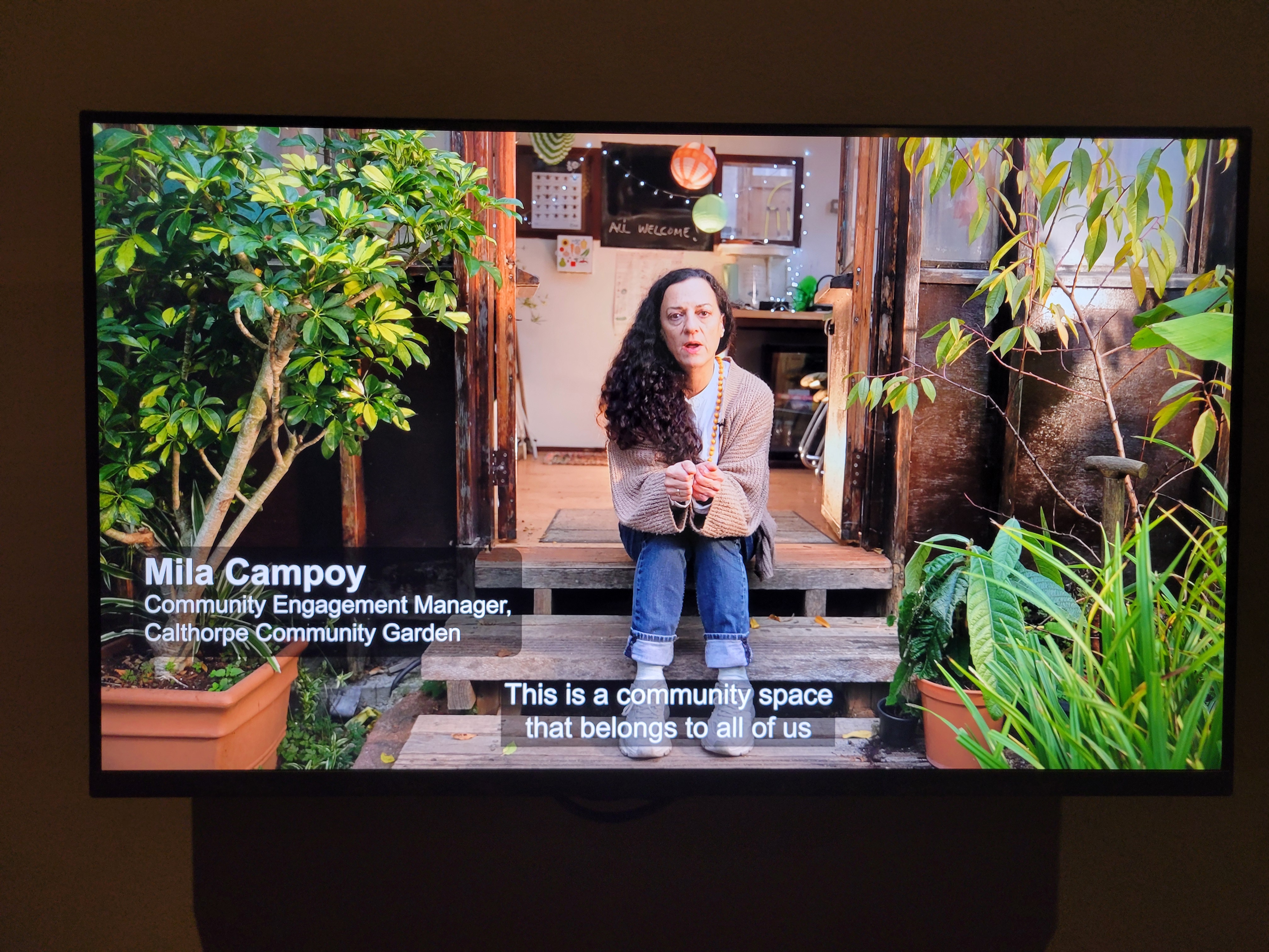 Mila from Cathorpe Community Garden sits on a step. Subtitles say 'this is a community space for all of us.'