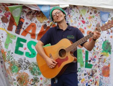A smiling person plays guitar in front of a handpainted sign saying London Harvest Festival