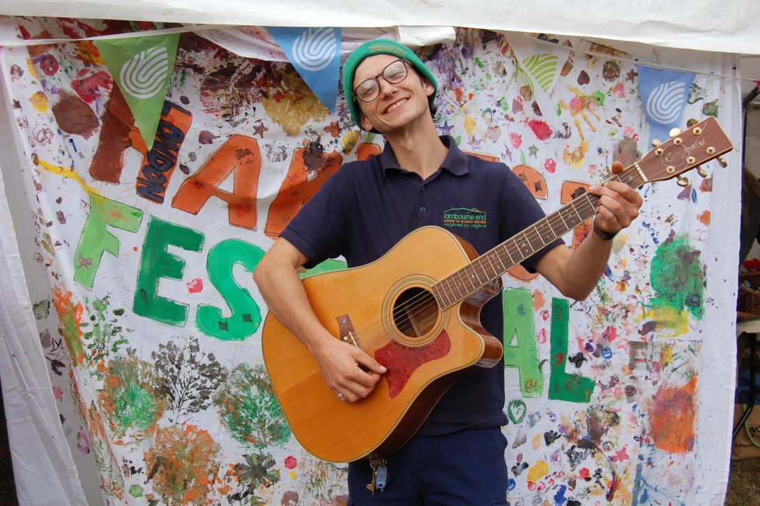 A smiling person plays guitar in front of a handpainted sign saying London Harvest Festival